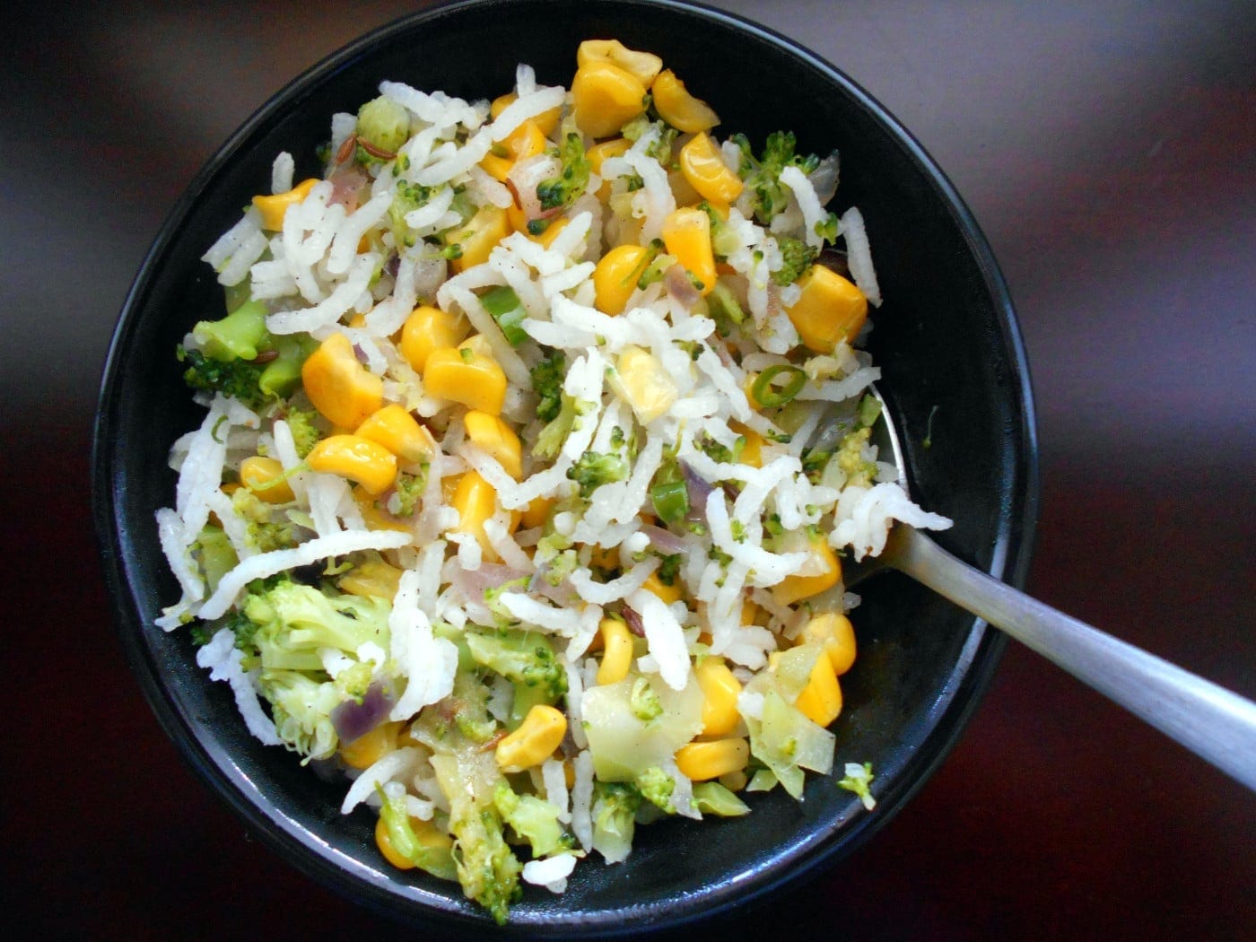 A small black bowl with rice, corn and broccoli with a spoon