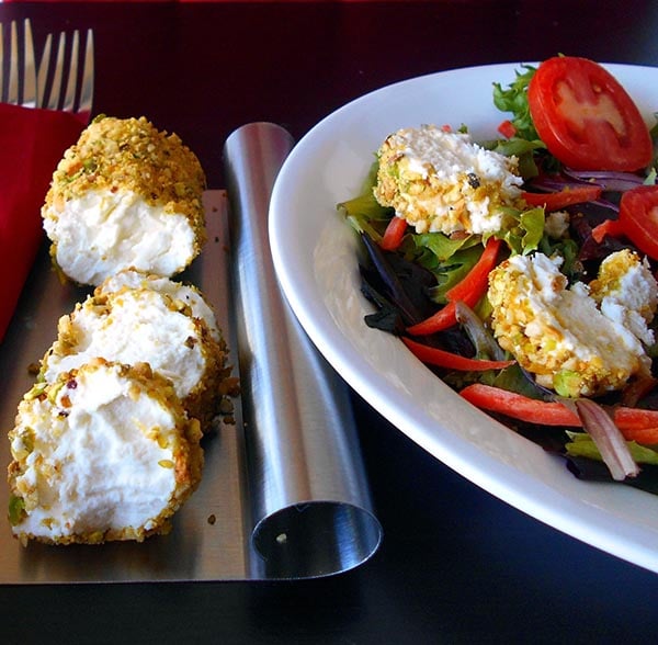 Front View of Goat cheese Salad log coated in pistachio and cardamom and cut into rounds. A salad plate with greens and goat cheese rounds is on the side