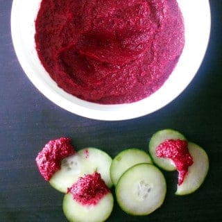 Top view of a white bowl with beet hummus. On the side, cucumber slices dipped in the hummus