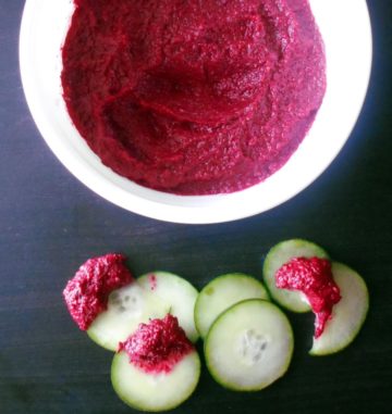 Top view of a white bowl with beet hummus. On the side, cucumber slices dipped in the hummus