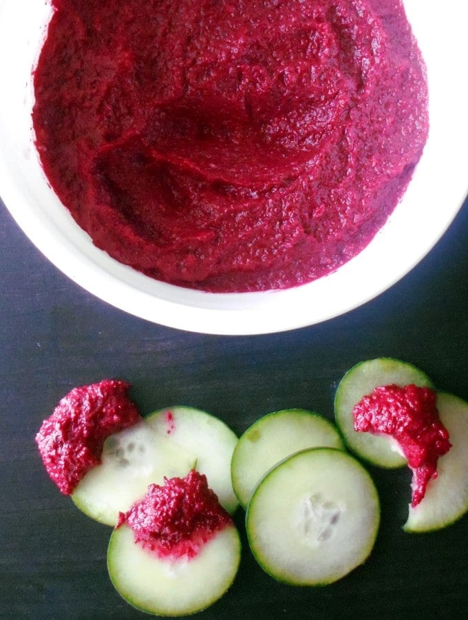 Top view of a white bowl with beet hummus. On the side, cucumber slices dipped in the hummus