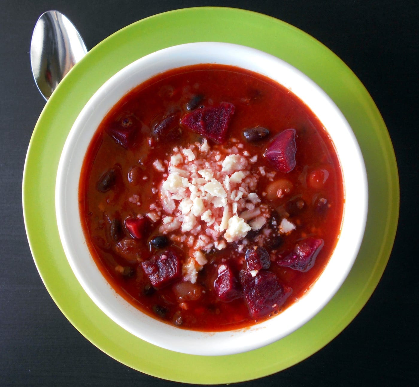 Overhead View of a White Bowl Filled with Vegetarian Beet Chili with Crumbled Romano Chili in the Middle of the Bowl. Bowl is on Top of a Light Green Plate. Top of a Silver Spoon is Visible on the Top Left Side of the Picture and It Is Under the Green Plate