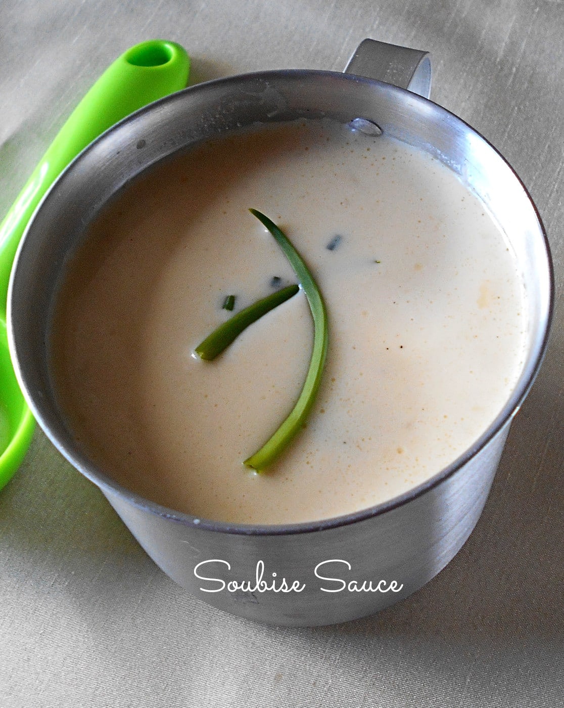 Front View of a Steel Measuring Cup Filled With Soubise Sauce and a Green Plastic Spoon Next to it