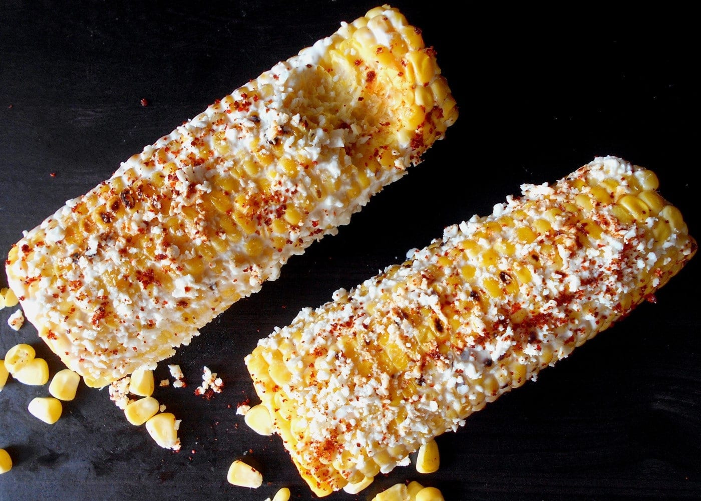 top view of 2 corn on the cobs topped with cheese and pepper.  One has a bite taken out of it.