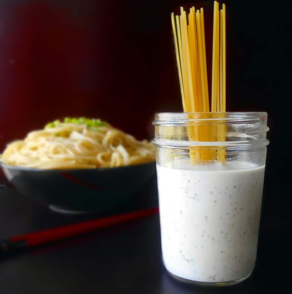 Front view of a mason jar filled with spicy lime dressing with spaghetti in it. In the back, a bowl of noodles is visible