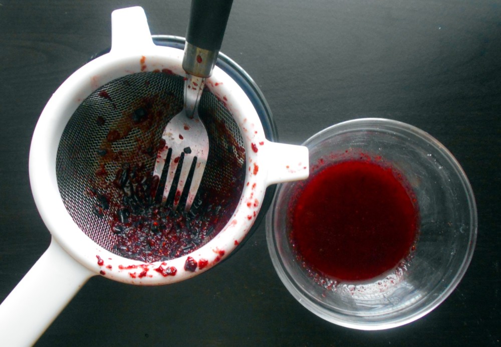 Overhead view of a sieve with blueberry pulp and a glass bowl on the side with blueberry juice