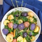 Overhead view of white bowl with a blue napkin wrapping most of the bowl. Bowl filled with multi colored potatoes, sautéed greens, carrot shavings, grilled shallots and pesto sauce.