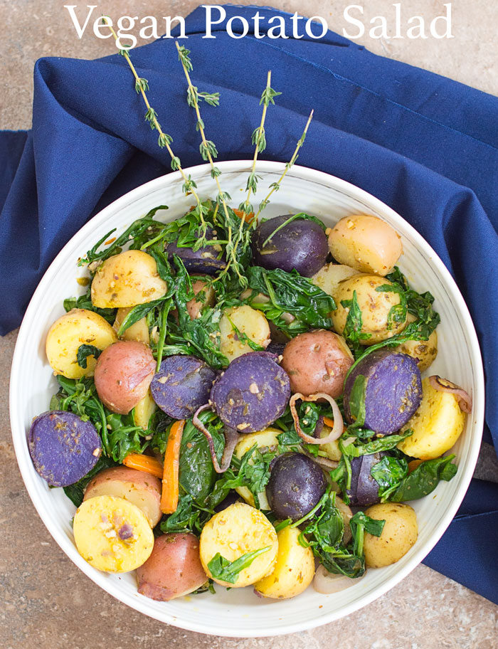 Overhead view of white bowl with a blue napkin wrapping most of the bowl. Bowl filled with multi colored potatoes, sautéed greens, carrot shavings, grilled shallots and pesto sauce.