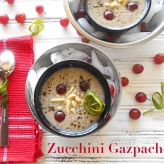 Overhead View of a Small Black Bowl with Cream Colored Zucchini Gazpacho Garnished with Grapes and Slivered Almonds. Black Bowl is on Top of a Slightly Larger Stainless Steel Bowl Filled With Ice Cubes. On the Left Side, a Spoon Sits On Top of a Light Red Napkin. On the Top Right Corner Is a Similar 2 Bowl Setup that is Partially Visible. A Few Grapes are Strewn About in the Picture. Zucchini Gazpacho is Written in Didot Font at the bottom of the Photo.