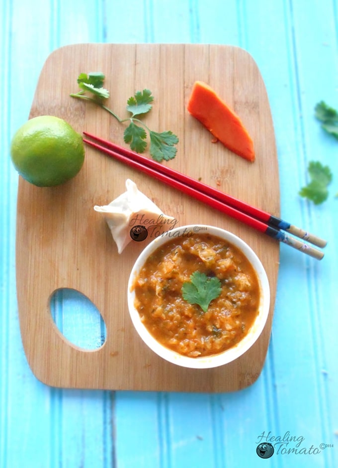 Overhead view of a chopping board on a blue surface. Chopping board has 1 lime, a slice of papaya, cilantro leaves, a samosa wonton, 2 chopsticks and a red bowl filled with papaya chutney