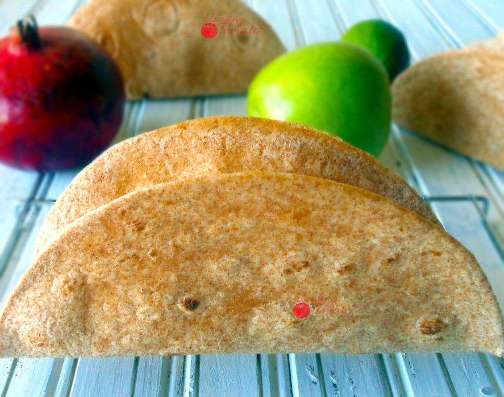 Front View of the Side of 1 Hard Taco Shell on a Cooling Rack with Pomegranate, Green Apple, Lime and 2 Taco Shells in the Background