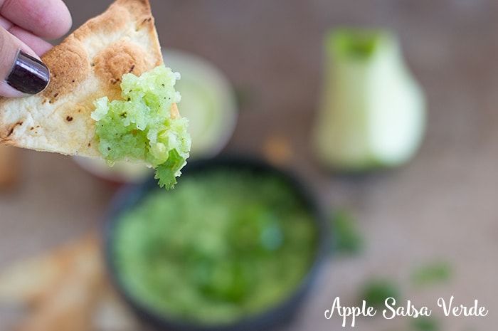 The author's fingers holding a chip filled with the apple salsa verde. In the back the bowl of salsa verde, apples and chips are blurred