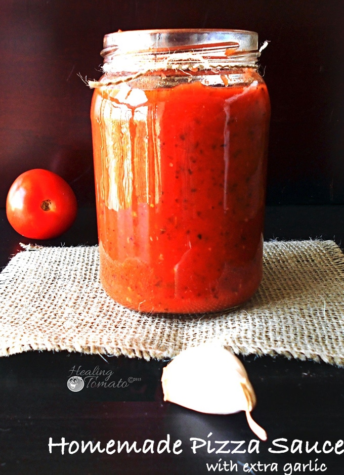 Closeup View of a Mason Jar Filled With Pizza Sauce. There is a Garlic Clove in Front of the Jar and a Plum Tomato in the Background