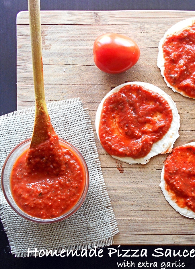 Overhead View of a bowl filled With Pizza Sauce and a Wooden Spoon in it. 3 Mini Uncooked Pizza Topped with the Sauce is on the Right. There is a Plum Tomato on the Top Right of the Photo