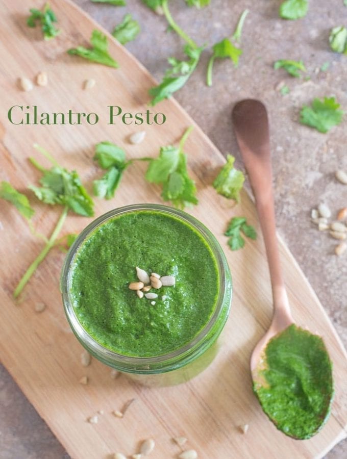 Overhead view of cilantro pesto in a mason jar with a brass spoon that had been dipped in the pesto sauce. A few leaves of cilantro scattered arond mason jar. A few sunflower seeds placed in the middle of the pesto sauce.
