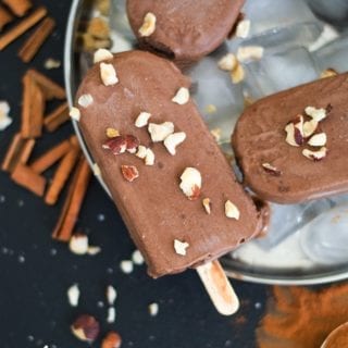 Overhead View of one Chocolate Popsicle Topped with Chopped Hazelnut. The Popsicle in on Top of Ice Cubes in a Steel Plate. 2 Other Partially Visible Popsicles are also placed on the Plate. The Plate is surrounded by Cinnamon Sticcks, Chopped Hazelnuts and Cinnamon Powder
