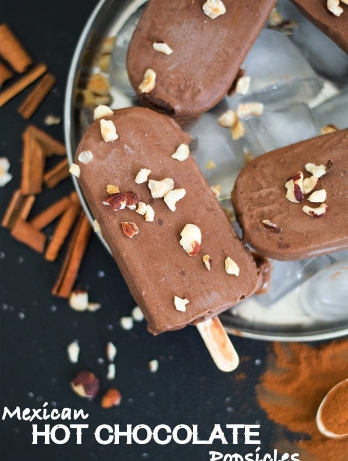 Overhead View of one Chocolate Popsicle Topped with Chopped Hazelnut. The Popsicle in on Top of Ice Cubes in a Steel Plate. 2 Other Partially Visible Popsicles are also placed on the Plate. The Plate is surrounded by Cinnamon Sticcks, Chopped Hazelnuts and Cinnamon Powder