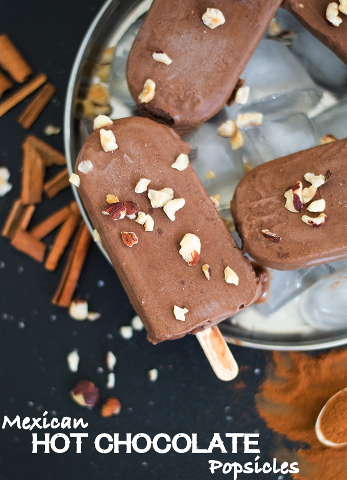 Overhead View of one Chocolate Popsicle Topped with Chopped Hazelnut. The Popsicle in on Top of Ice Cubes in a Steel Plate. 2 Other Partially Visible Popsicles are also placed on the Plate. The Plate is surrounded by Cinnamon Sticcks, Chopped Hazelnuts and Cinnamon Powder