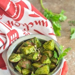 Roasted Potatoes in Cilantro Pesto Viewed From a 75° Angle. Roasted Potatoes are in a Copper Bowl Under a white and Orange Kitchen Towel with Sprigs of Cilantro in the Distant Background