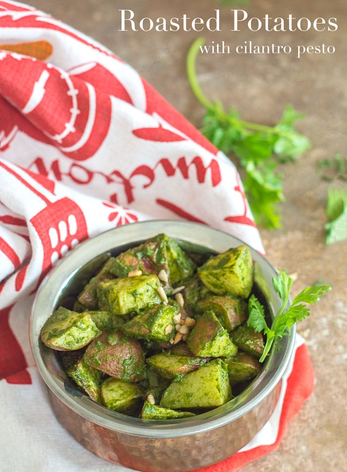 Roasted Potatoes in Cilantro Pesto Viewed From a 75° Angle. Roasted Potatoes are in a Copper Bowl Under a white and Orange Kitchen Towel with Sprigs of Cilantro in the Distant Background