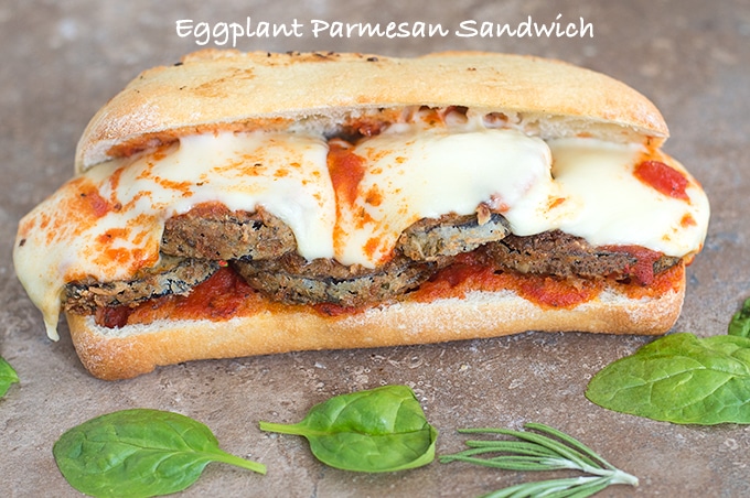 Front View of an Eggplant Parmesan Sandwich with Melted Mozzarella Cheese and Surrounded by Spinach Leaves and Rosemary Sprigs