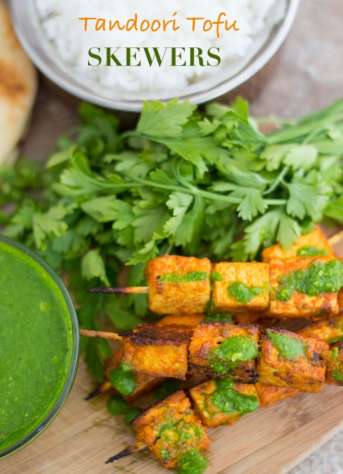 Overhead View of Skewered Tofu Oven Roasted and Stacked on each other. Two Skewers with 3 Tandoori Tofus Each are Placed at the Bottom of the Stack and on Top of a Brown Board. A Green Chutney is Lightly Drizzled on Top. Two Other Skewers With four 1” Tofu Squares is Stacked on Top and Also Has Green Chutney Lightly Drizzled. A Bunch of Italian Parsley is Placed Behind the Skewers. Behind the Parsley, a Copper Bowl Filled with Rice is Partially Visible. Next to the Rice Bowl, There is a Naan That is Barely Visible. To the Left of the Skewers, There is a Glass Bowl with Green Chutney that is Partially Visible.