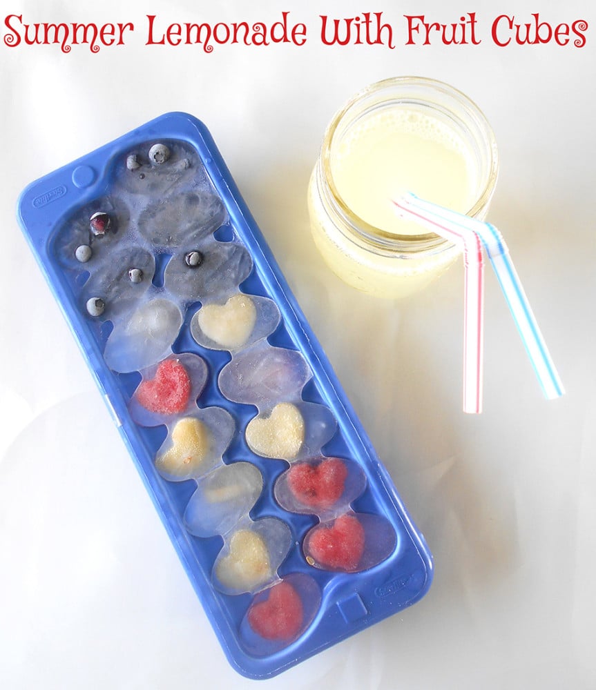 Overhead view of an ice cube tray with frozen ice cubes with fruits. A glass of lemonade is on the side