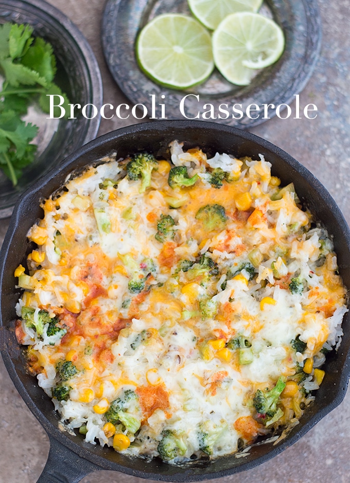 Overhead View of a Black Skillet Filled with Broccoli Rice Casserole and Topped with Cheese. Two Small Rustic Plates are Set on the Top Left. One is Filled with Lime Rounds and the Other is Filled with Cilantro Stems