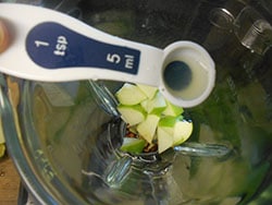 Overhead View of 1 Tbsp Measuring Spoon Filled With Lime Juice Hovering Over Chopped Apples and Hazelnuts Inside a Blender
