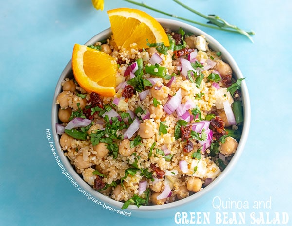 Closeup view of a bowl on a blue background. Bowl is filled with quinoa, green beans and chick peas. Garnished with cilantro and orange slices. Small yellow carnations on the side - Green Bean Salad
