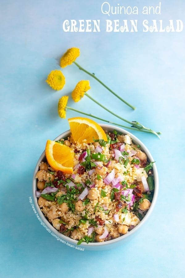 Overhead view of a bowl on a blue background. Bowl is filled with quinoa, green beans and chick peas. Garnished with cilantro and orange slices. Small yellow carnations on the side - Green Bean Salad