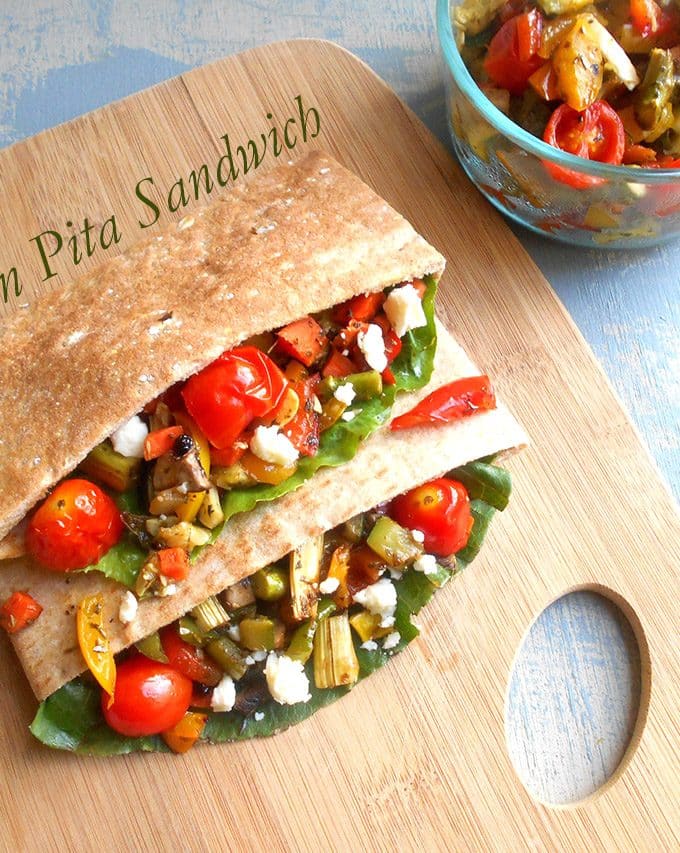 Overhead view of 2 Pita Sandwiches Stuffed with Roasted Veggies Sitting on a Chopping Board and a Glass Bowl with Roasted Veggies on the Top Right Side of the Picture.