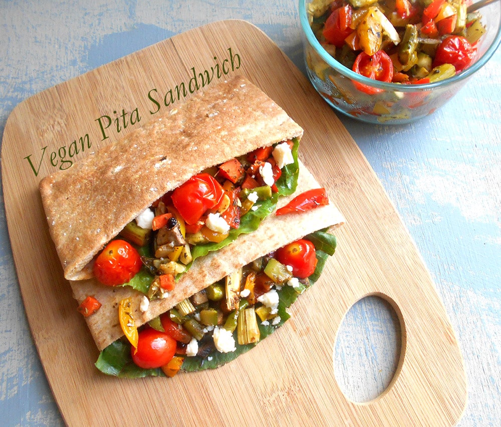 Overhead view of 2 Pita Sandwiches Stuffed with Roasted Veggies Sitting on a Chopping Board and a Glass Bowl with Roasted Veggies on the Top Right Side of the Picture.