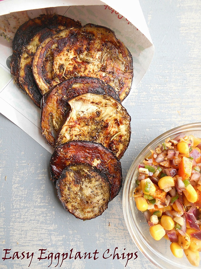 Overhead View of Eggplant chips laying flat on a blue board. They are arranged in a fan shape coming out of a newspaper cone. On the bottom right of the photo, a small glas bowl with corn salsa is visible