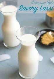 Two Small Glass Vases Filled with Lassi. One is in the Foreground and the Other is in the Back to the Left. 3 Ice Cubes Strewn About. In the Background to the Right, There is a Tiny Black Dish Filled with Cumin Powder. Salted Lassi