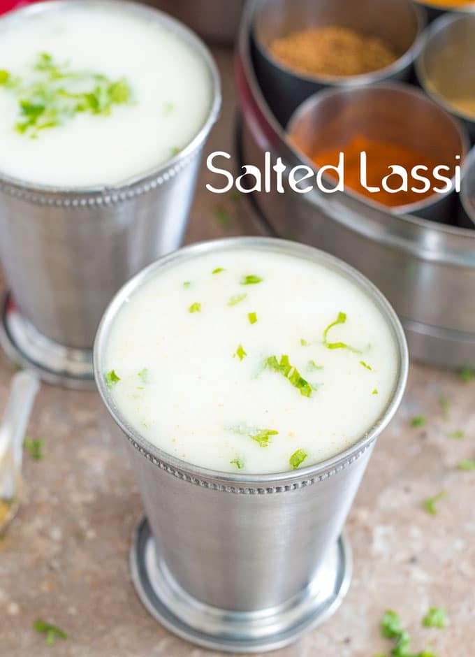 Closeup and 75° view of a stainless steel Mint Julep Cup Filled with White Liquid Filled to the Brim and Garnished with Chopped Cilantro. To the Left Background, There is a Similar Stainless Steel Cup Filled with to the Brim with the Same White Liquid and Garnished with Chopped Cilantro. To the Right Background, there is an Circular Indian Stainless Steel Container that is Partially Visible. It is filled with Small Cyndrical Containers Arranged in a Circular Pattern. Each Container Contains Different Indian Spices. There is Chopped Cilantro Strewn All Over the Brown Tile on which All the Items are Placed. Salted Lassi