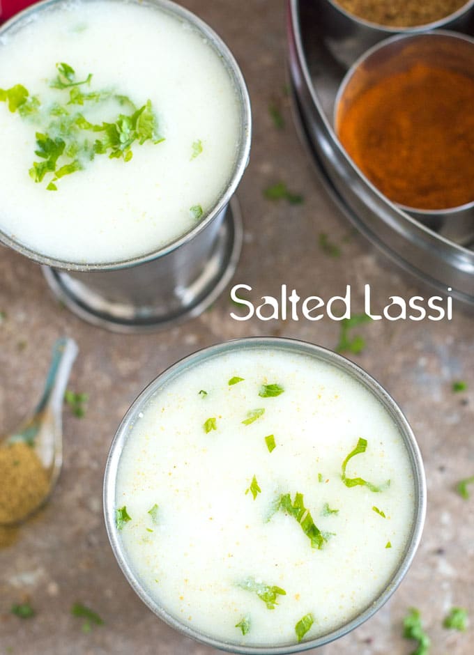 Salted Lassi. Overhead View of 2 Stainless Steel Mint Julep Cup Filled with White Liquid Filled to the Brim and Garnished with Chopped Cilantro. One cup is at the Bottom of the Photo and the Other is on the Top Left. On the Top Right, There is an Circular Indian Stainless Steel Container that is Partially Visible. It is filled with Small Cyndrical Containers Arranged in a Circular Pattern. Each Container Contains Different Indian Spices. There is Chopped Cilantro Strewn All Over the Brown Tile on which All the Items are Placed. There is a Tiny Stainless Steel Spoon on the Bottom Left in the Photo Filled with Cumin Powder.