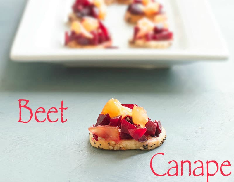 One bagel chip on a blue board and topped with beetroot and pineapples. In the back, there are bagel chips on a white plate and topped with beetroot and pineapples