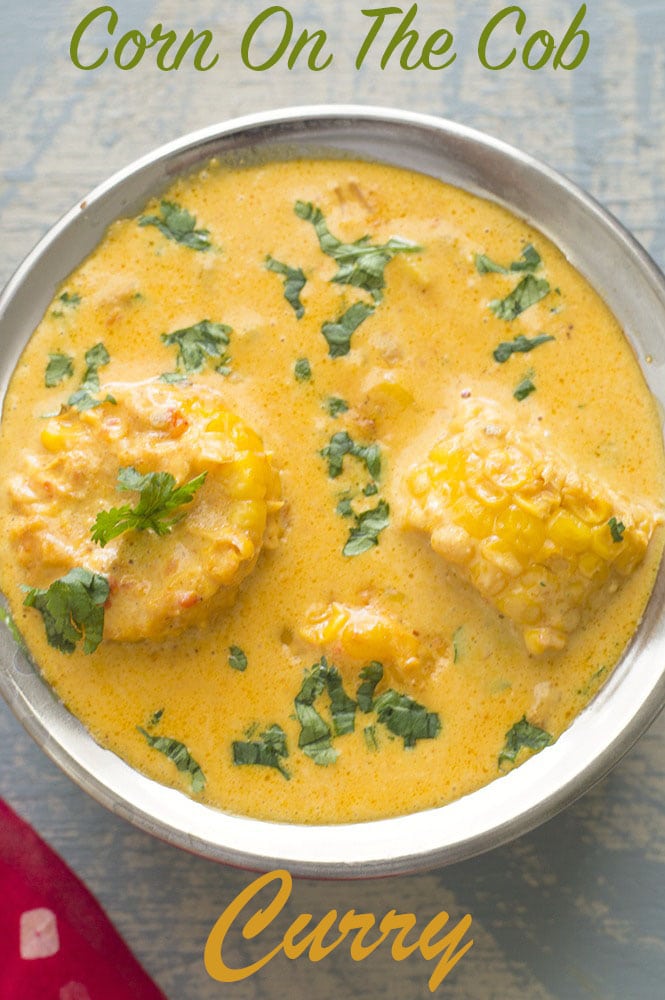 Overhead View of a Copper Bowl Filled with Yellow Curry. Two Pieces of Corn on the Cob Potruding from the Curry and Chopped Cilantro Strewn on the Surface of the Curry as Garnish. A Small Piece of a Red Scarf with White Dots is Seen on the Bottom Left of the Photo