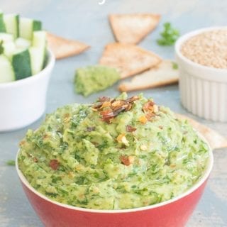 Front View of a Red Bowl Filled with Parsley Hummus and Topped with Pepper Flakes. The Hummus Top is Shaped like a Dome. On the Top Right, there is a White Ramekin Filled with Sesame Seeds. On the Top Right, there is a Ramekin Filled with Cucumber Pieces. There are 3 Pita Chips Placed Around the Bowl