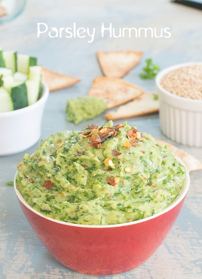 Front View of a Red Bowl Filled with Parsley Hummus and Topped with Pepper Flakes. The Hummus Top is Shaped like a Dome. On the Top Right, there is a White Ramekin Filled with Sesame Seeds. On the Top Right, there is a Ramekin Filled with Cucumber Pieces. There are 3 Pita Chips Placed Around the Bowl