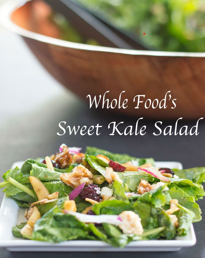 Front View of a White Plate With Kale, Cranberries, Dried Apples, Walnuts, Red Onions, Carrots, Sunflower Seeds, Sliced Almonds, Feta and Orange Zest. Background has a large Brown Salad Bowl That is Delibrately Blurred. Contains Kale Salad With Thin Black Plastic Tongs.