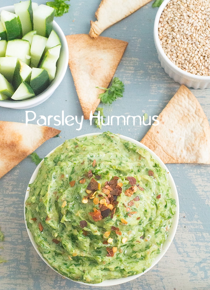 Overhead View of a Red Bowl Filled with Parsley Hummus and Topped with Pepper Flakes. On the Top Right, there is a White Ramekin Filled with Sesame Seeds. On the Top Right, there is a Ramekin Filled with Cucumber Pieces. There are 3 Pita Chips Placed Around the Bowl