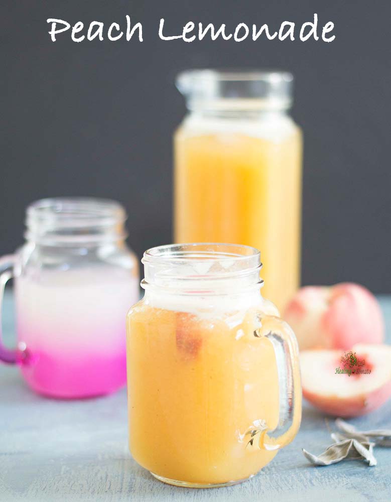 Front view of a glass mug with peach lemonade. Peaches and sage on the side and a pitcher of peach lemonade in the back. 