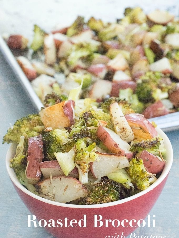 Front View of a Small Red Bowl Overflowing with Roasted Broccoli and Potatoes. In the Background, a Baking Tray Filled with Roasted Broccoli and Potatoes is Blurred Slightly