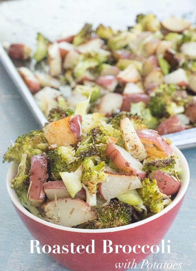 Front View of a Small Red Bowl Overflowing with Roasted Broccoli and Potatoes. In the Background, a Baking Tray Filled with Roasted Broccoli and Potatoes