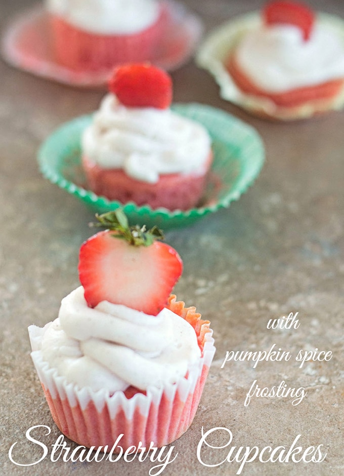 Front View of a Strawberry Cupcake With Frosting and a Sliced Strawberry. In the Background, there are 3 More Cupcakes