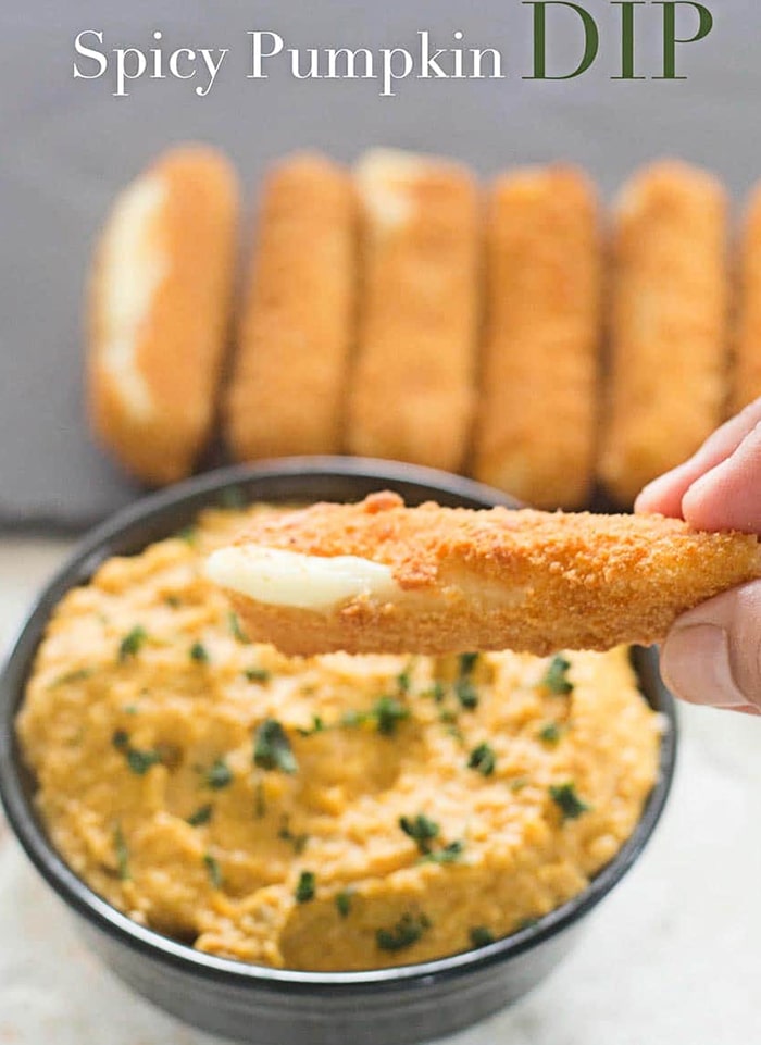 Front View of the Author’s Fingers Holding a Baked Mozzarella Stick Oozing With a Little Cheese. Mozzarella Stick is Hovering Over a Black Bowl with Yellow Spicy Pumpkin Dip Garnished with Chopped Curley Parsley. In the Background, 4 Sticks of Baked Mozzarella with Cheese Oozing Out in Two of Them. The Mozzarella Sticks are on an Grey Basalt Matt Serving Tray