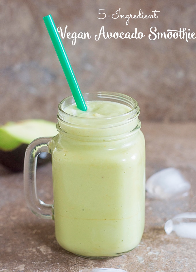 Front View of a Smoothie Glass Filled with the Avocado Smoothie. Ice Cubes Strewn About and Half of an Avocado Visible in the Background