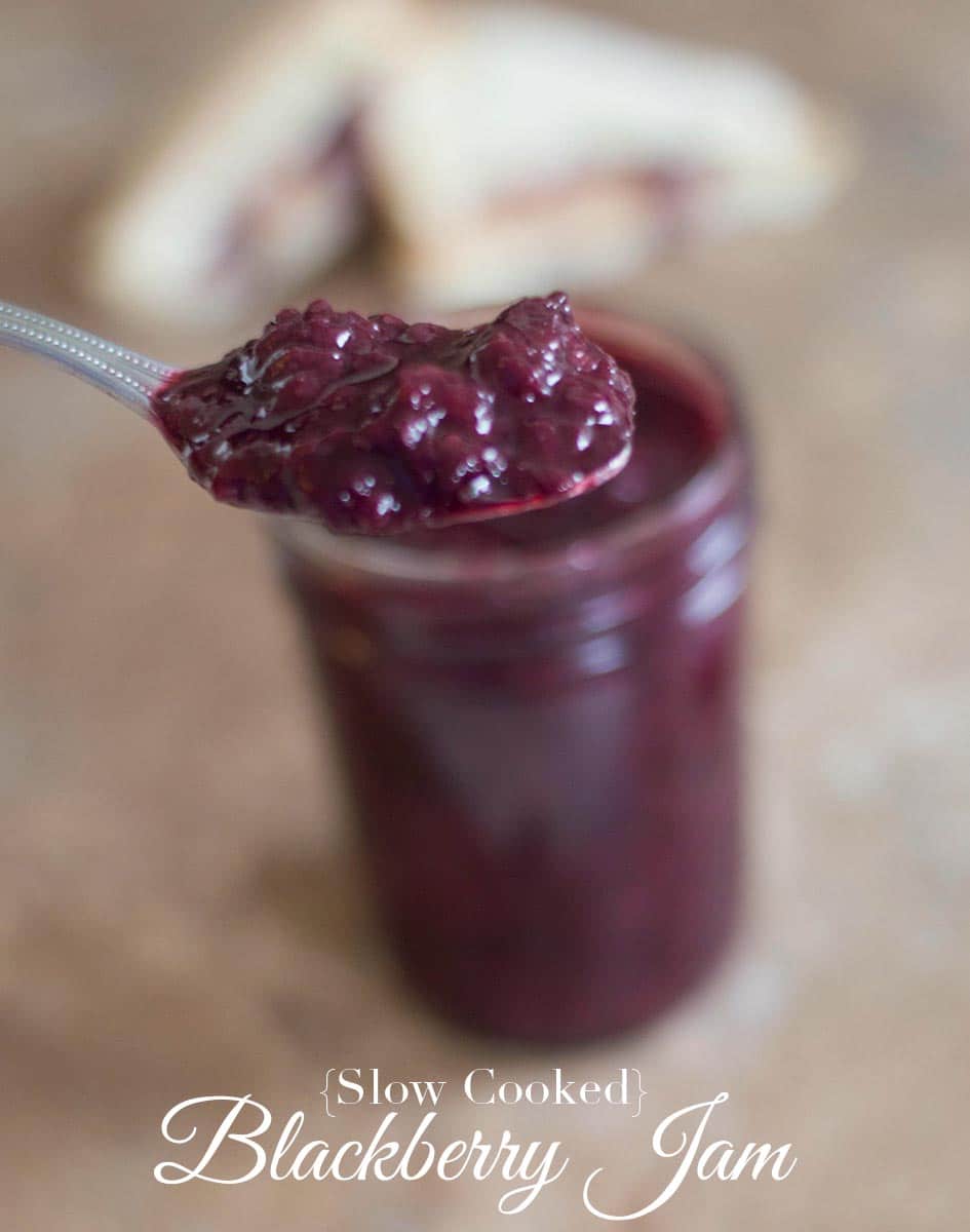 Front fiew of a spoon filled with blackberry jam hovering over a mason jar filled with the same jam. In the back 2 small pb&j recipes are stacked on top of each other